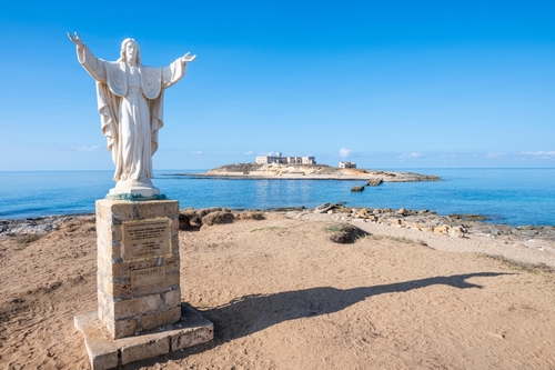 The statue of Christ Redeemer in Portopalo with the Island of the currents (isola delle Correnti) on the Ionian coast in the background, located in the territory of the municipality of Portopalo, Sicily, Italy