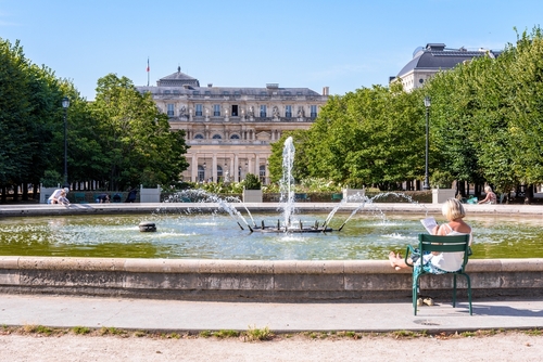 A senior woman is relaxing with a word search puzzle, sitting on a metal lawn chair by the fountain in the Palais-Royal garden on a sunny summer morning, Paris, France