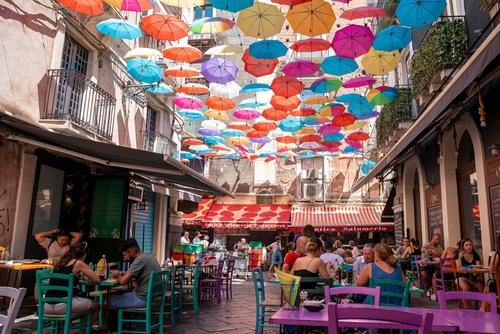 Male and female tourists relaxing at cafe under colorful umbrellas hanging amidst buildings. View of beautiful decoration on streets in city during summer. Catalina, Sicily, Italy