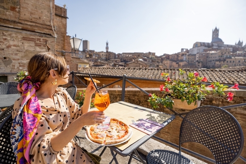 Young woman having lunch with pizza and wine at outdoor restaurant with beautiful view on the old town of Siena, Tuscany, Italy. Concept of Italian cuisine and traveling in Tuscany