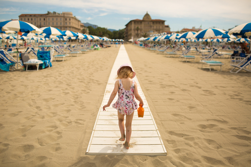 A little girl walks on a path on the beach of Viareggio, Lucca, Tuscany, Italy. In front of it there are beach umbrellas and city buildings