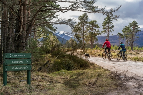 Path to the Lairig Ghru though the Cairngorms National Park with local signage in foreground and cyclists with dog heading towards the mountains under a cloudy sky, Aviemore, Scotland, UK