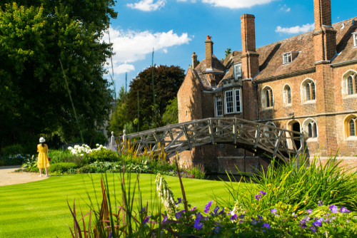 Girl in yellow dress stands on public park enjoying the picturesque Mathematical Bridge and the public Old Library at Cambridge, England, UK