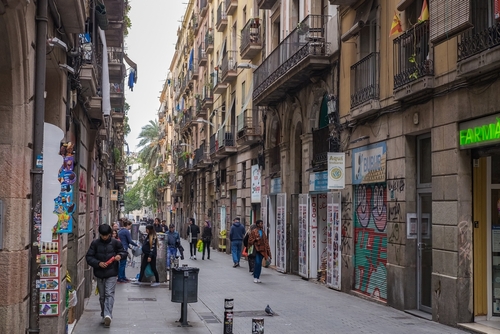 Street located in the Raval neighborhood, showing people walking, shops, apartments balconies and a bunch of palm trees in the background in Barcelona, Catalonia, Spain