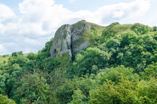 View of Thor's Cave with blue sky and green trees all around in the Peak District National Park, Derbyshire, England, United Kingdom