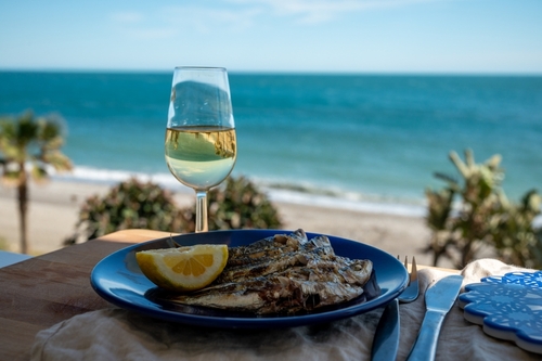 Sardines espeto prepared on skewers and open flame on fireplace with olive trees wood, served outdoor with lemon and glass of fino sherry wine and view on blue sea, Malaga, Andalusia, Spain