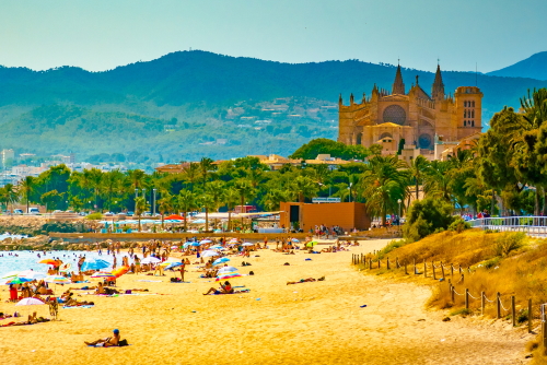 View of the beach of Palma de Mallorca with people lying on sand and the gorgeous cathedral building visible in the background, Mallorca island, Balearic islands, Spain