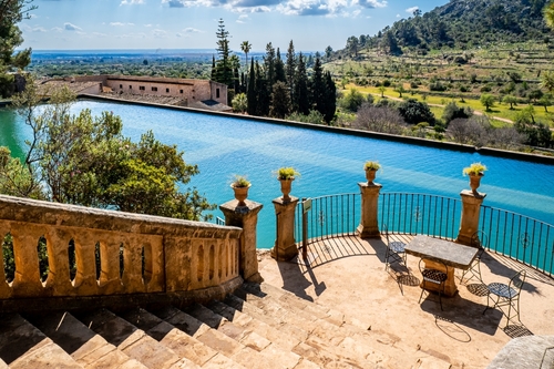View downstairs over a semicircular terrace in front of a water basin with planted amphorae, a stonemade table and four iron chairs above the vernal gardens of a public estate called Raixa on Mallorca Island, Balearic Islands, Spain