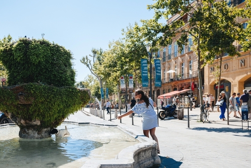 People pass leisurely in the hot sun of Cours Mirabeau, Aix-en-Provence. Trees and The Fountain des Neufs Canons grace a unique town plaza, Provence, France