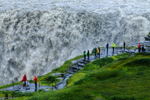 Tourists at a wet and powerful Dettifoss waterfall as it gushes into Jökulsárgljúfur canyon, with rising mist and spray and green Summer grasses, Vatnajökull National Park, North Iceland