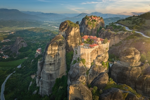 Aerial view of Meteora Monastery complex on the rocks, illuminated by rising sun against mountains in background, a UNESCO World Heritage Site in Greece