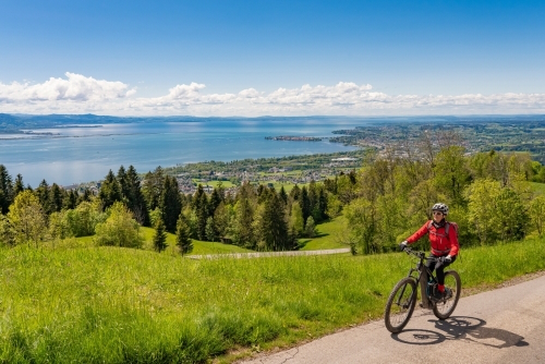 Happy, active senior woman cycling above Lake Constance, Bodensee and admiring the awesome view over Lake Constance to city of Lindau in background, Germany