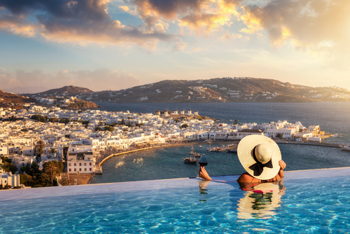 A woman with a glass of wine, bathing in a swimming pool, enjoying the view over the town of Mykonos on Mykonos Island, Cyclades Islands, Greece, during a Summer Sunset