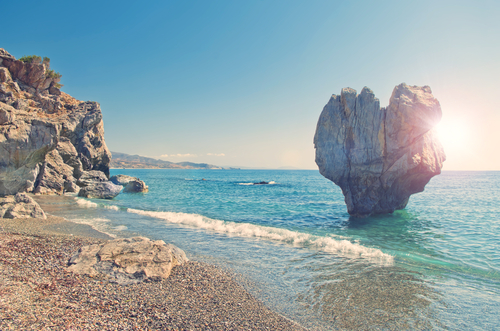 View of a huge heart shaped rock on pebble beach of Preveli with sun shining behind the rock in Rethymno, Crete island, Greece