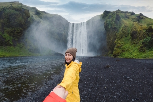 Asian woman in yellow jacket holding hands with couple at front of Skógafoss waterfall flowing on cliff in Summer, Landmannalaugar Reservation, Iceland