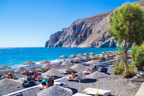 View of the Black Beach of Kamari village and its facilities, people relaxing on the beach at the foot of Mesa Vouno mountain, Santorini Island, South Cyclades Islands, Greece