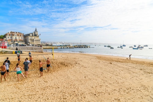 A group of active young adult men and women play a game of beach volleyball in the sand at the Praia da Ribeira beach in the picturesque seaside town of Cascais, Portugal